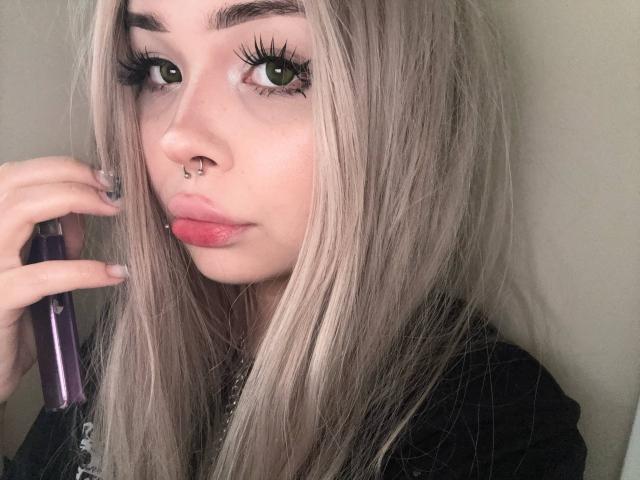 SerinaSindlinger with long blonde hair, green eyes, black eyeliner, pink lipstick, nose ring, holding a purple phone, wearing a black shirt, looking at camera with slightly parted lips.