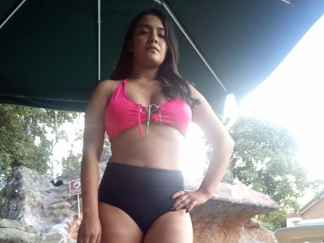 ArianithaXox, con senos de tamaño medio, lleva un top de bikini rosa con cordones y tanga negra de cintura alta, está de pie debajo de un paraguas verde, mirando hacia abajo con una expresión seria..