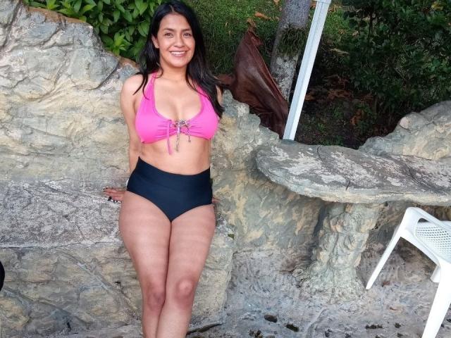 ArianithaXox, con senos de tamaño medio, con un top de bikini rosa y pantalones negros de cintura alta, de pie afuera, sonriendo, con los brazos detrás de la espalda, frente a un terreno rocoso, con una silla blanca..
