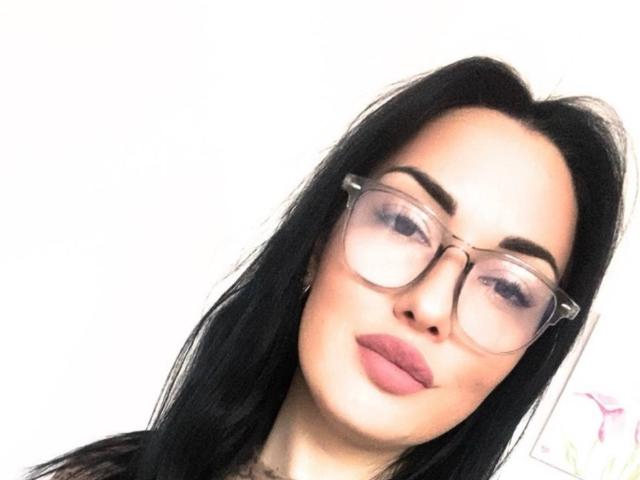 Xioni has black hair, clear glasses, and pink lipstick. She's looking sideways with a slight smile. Her ass is not visible. She has a fair skin tone.
