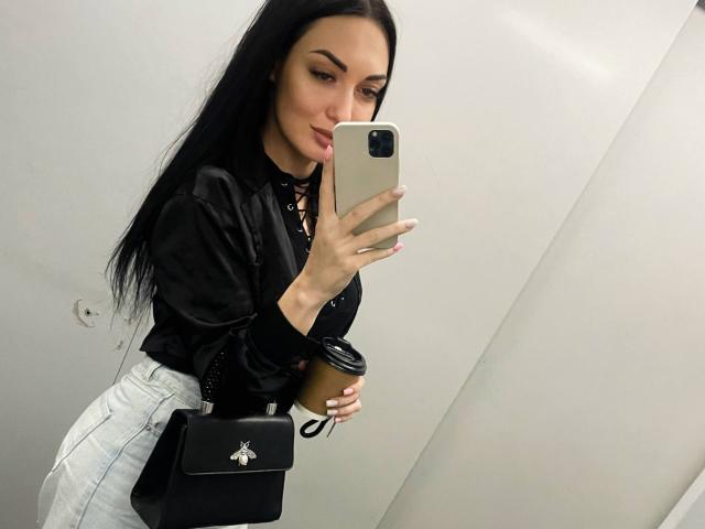 Xioni with long black hair, black lace-up shirt, white high-waisted jeans, black purse, holding coffee cup, taking selfie, pouting, phone in hand, white wall in background.