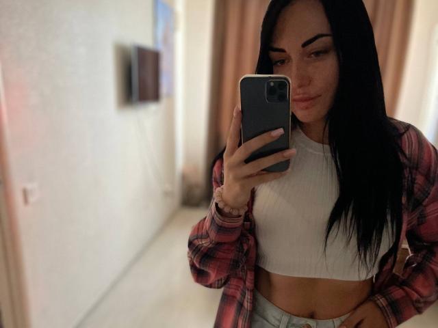 Xioni with long black hair, wearing a red plaid shirt and white ribbed crop top, holding a phone, reflecting in a mirror, showing her flat stomach and small tits.