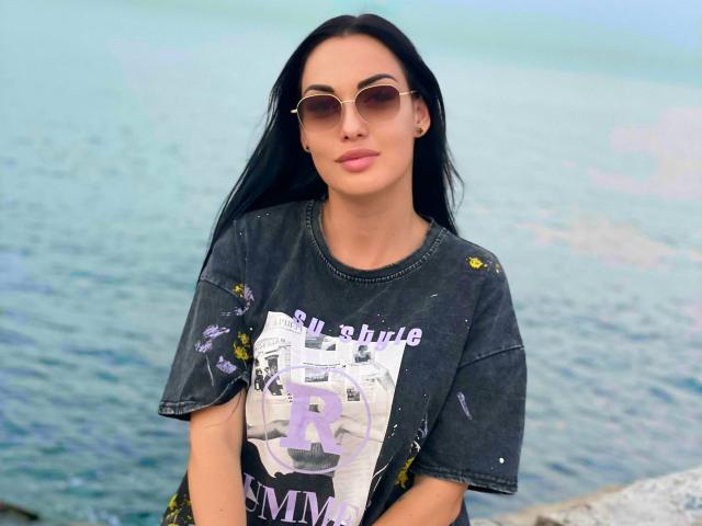 Xioni with long black hair, wearing a dark tie-dye shirt with purple lettering, and round sunglasses, standing by a blue water body, has a cute, confident look.