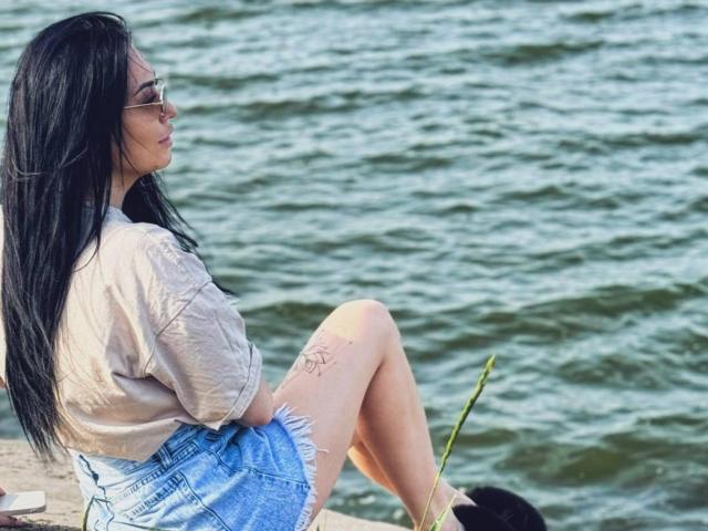 Xioni has long black hair, wears glasses, a beige shirt, and blue denim shorts, sitting by the water with a tattoo on her right thigh and a grass stem.