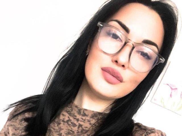 Xioni has black hair, clear glasses, and pink lips. She's wearing a brown lace top. Her ass is not visible, and her pussy is not shown. She's smiling slightly.