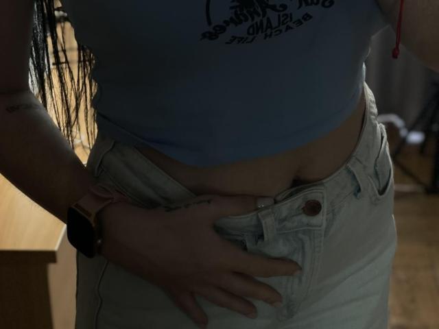 Xioni's blue crop top, pale skin, and white jeans, with her hand on her exposed belly button, and her pink wristwatch visible, in a dimly lit room.