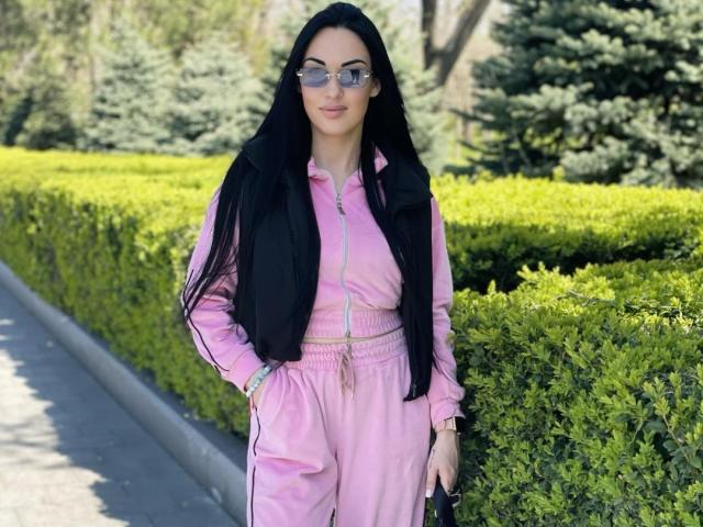 Xioni has long black hair, wearing a pink track suit with a black vest, and sunglasses, standing on a sidewalk with green bushes and trees in the background.