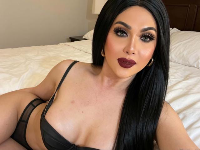 EhlaGrey with long black hair, dark lipstick, and black bra, lying on a bed, showing her tits and looking at the camera with thick, curly eyelashes.