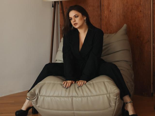 Dreia, sentada em um banquinho de couro bege, vestindo um vestido preto com um decote profundo em V, saltos altos pretos e brincos, olhando sedutoramente para a câmera..