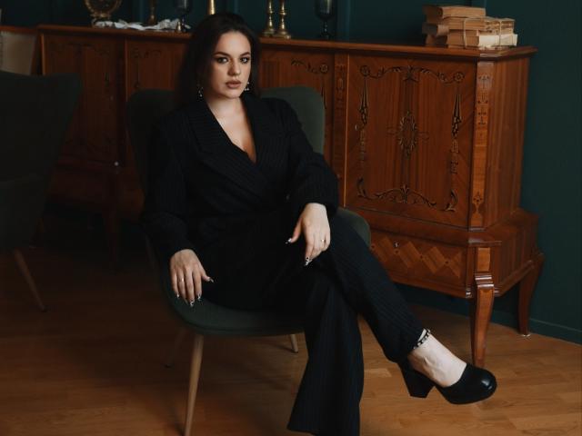 Dreia with black pinstripe suit, black heels, and medium tits, sitting in green chair, wooden cabinet in background, dark green wall, hardwood floor, looking serious, dark hair.