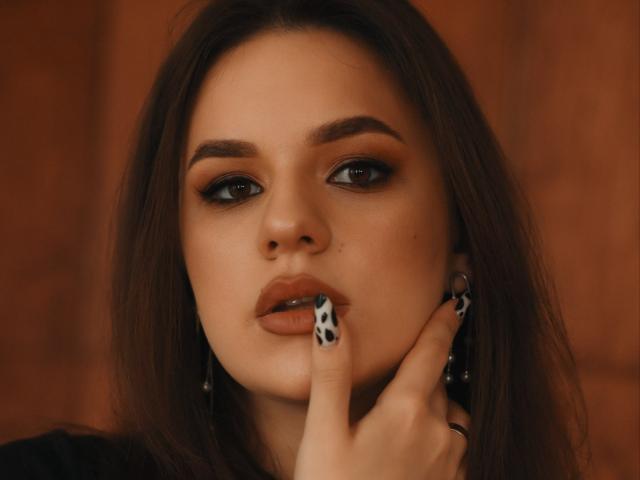 Dreia, met bruin haar, donkere oogmake-up en bruine lippen, houdt haar vinger tegen haar licht geopende, glanzende bruine lippen. Ze draagt zwarte oorbellen en haar vinger heeft een koeienprint op de nagel..