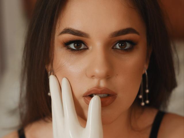 Dreia's face with brown eyes, dark brown hair, and nude lips, touching white fingers to her mouth. She's wearing black earrings. Soft, warm lighting. She has a small mole.