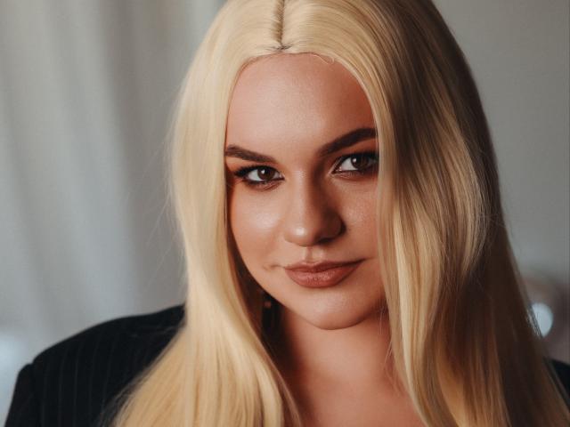 Blonde Dreia with straight hair, brown eyes, and subtle makeup, smiling slightly, wearing a black top, has smooth skin, and a cute face, looking directly at the camera.