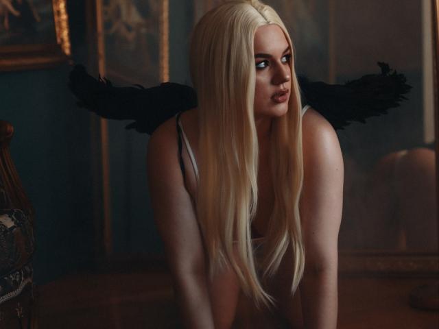 Blond-haired Dreia with black angel wings, leaning forward, showing off her ass in a low-cut top, with a seductive look in her eyes, in a dimly lit room.