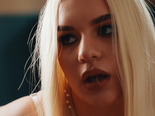 Dreia heeft blond haar, donkere ogen en lichtjes open lippen, waarbij een klein stukje van haar tong zichtbaar is. Ze draagt een parelketting en kijkt weg van de camera met een verleidelijke uitdrukking..