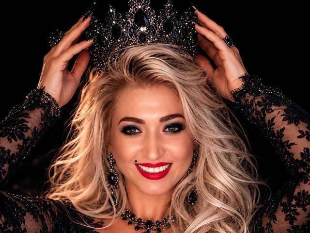 ElizabethRaegan, avec ses cheveux blonds, portant une robe noire en dentelle, une couronne, et du rouge à lèvres, souriante, tenant sa couronne avec ses mains, avec un vernis à ongles noir, un maquillage des yeux foncé, et des boucles d'oreilles chandelier noires..