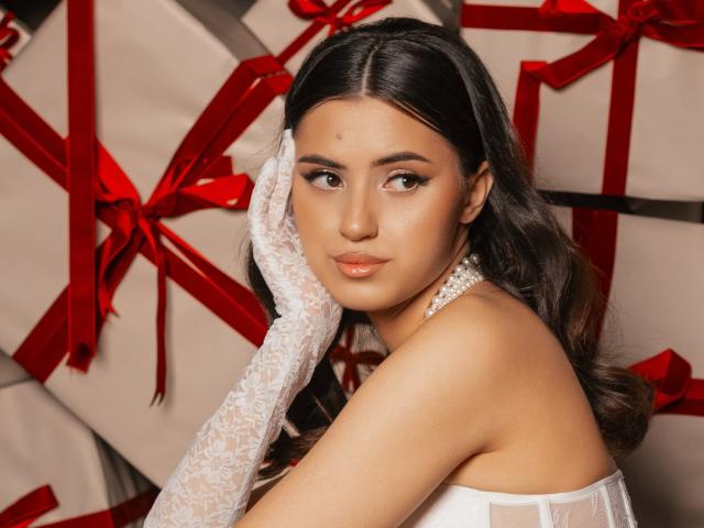 JasmineDay with long dark hair, wearing white lace gloves, touching her face, looking at the camera, wearing a white dress, surrounded by red ribbons and white boxes.