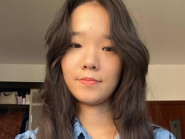 SamanthaTango a les cheveux longs mi-longs, ondulés et d'un brun foncé, et elle regarde directement l'appareil photo avec un sourire subtil et détendu. Elle porte une chemise bleue à motifs..