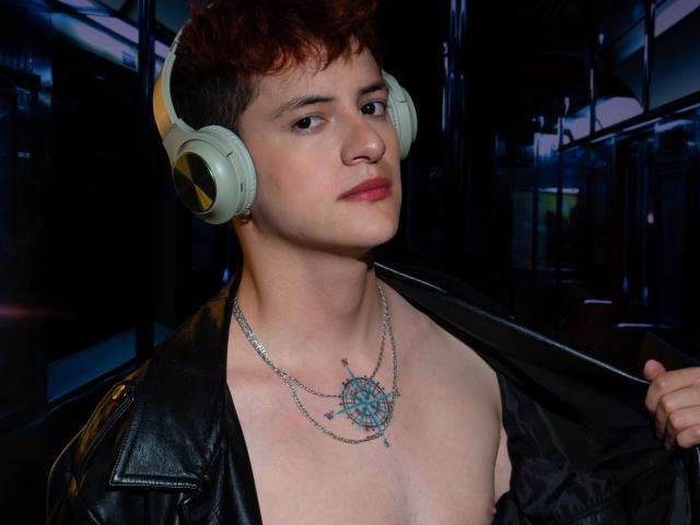 GabyThoms with short red hair, wearing beige headphones, black leather jacket, and a silver chain necklace with a blue gem, standing indoors, looking to the side, slightly smiling.