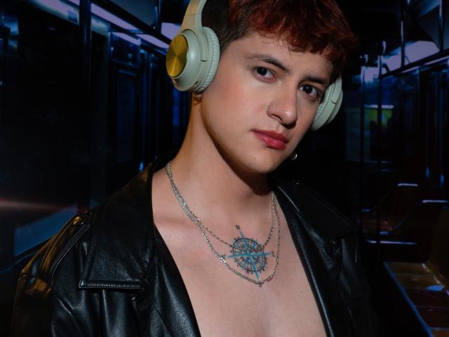 GabyThoms with short red hair, wearing a black leather jacket, silver necklace with a blue pendant, and beige headphones, looking slightly to the side with a neutral expression.