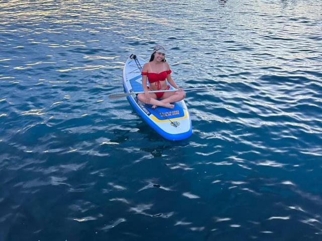 AmelyTorres with big tits, wearing a red bikini, sitting cross-legged on a blue and white paddleboard in dark blue water, looking relaxed.