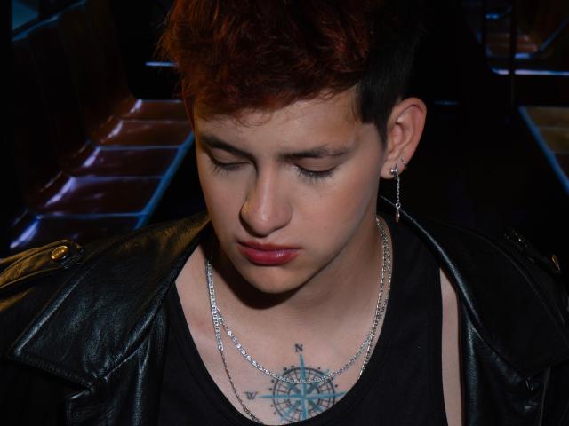 GabyThoms, met kort roodbruin haar, een zwarte leren jas en een ketting, kijkt naar een kompas-tatoeage op hun borst. Ze heeft roze lippenstift en oor piercings..
