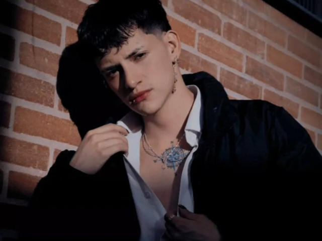 GabyThoms con i capelli neri corti, indossa una giacca nera, una camicia bianca e una collana con un ciondolo blu, tiene sollevato il colletto della giacca, ha un tatuaggio sul petto, espressione seria, muro di mattoni..