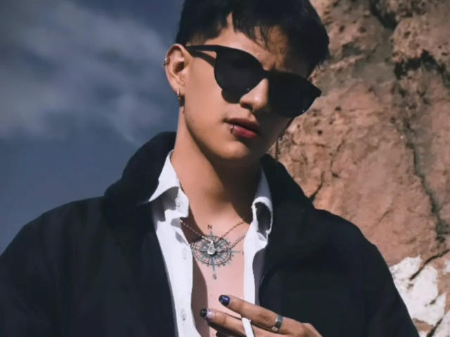 GabyThoms mit kurzen, schwarzen Haaren, schwarzen Sonnenbrillen, einem weißen Hemd, einer schwarzen Kapuzenjacke, der Sonne im Gesicht, einer Halskette, einem Ring und einer Hand, steht vor einer felsigen Wand..