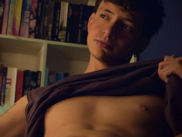 GabyThoms, con la camicia sollevata, che mostra addominali scolpiti e piccoli capezzoli, capelli castani, un leggero sorriso, una libreria sullo sfondo, illuminazione soffusa..