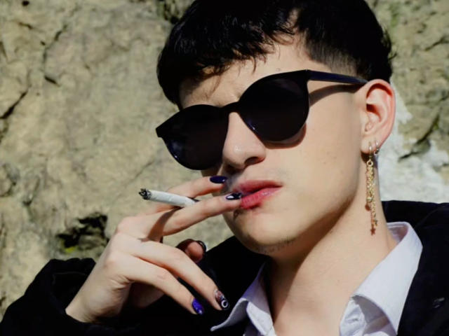 GabyThoms, avec des lunettes de soleil noires, tenant une cigarette entre ses lèvres ornées d'ongles violets, portant une chemise blanche et une veste noire, des boucles d'oreilles rock, des cheveux courts et foncés, devant un mur de pierre rugueuse..