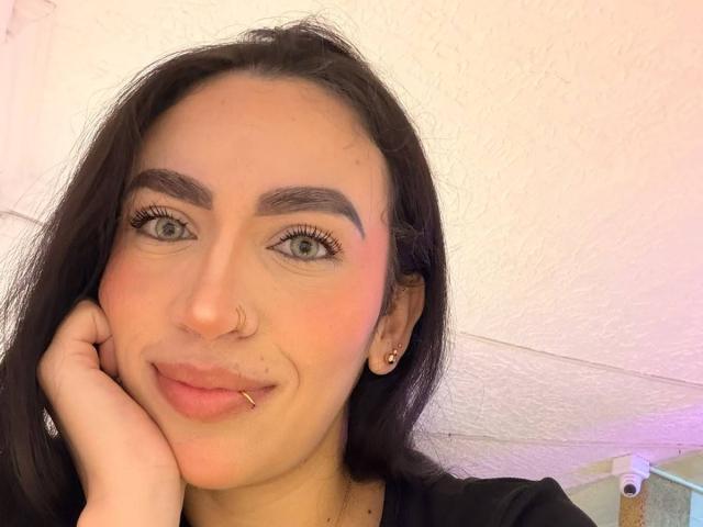 Camille, aspetto "JuicyHot", con capelli scuri, occhi verdi, guance rosate, piercing al naso e alle labbra, e orecchini, sorridente con una mano sul viso. Illuminazione rosa e bianca tenue..