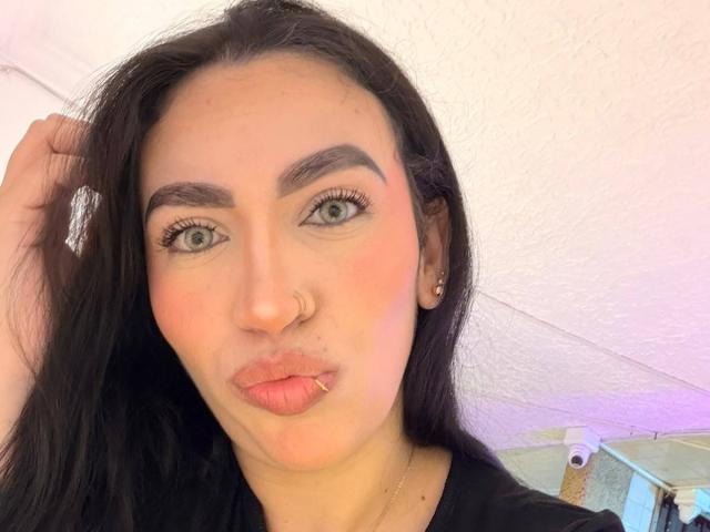 CamilleJuicyHot tem cabelo escuro, olhos verdes, um piercing no nariz, lábio inferior perfurado e um rosto fofo e expressivo, vestindo uma blusa preta, com uma mão no cabelo..