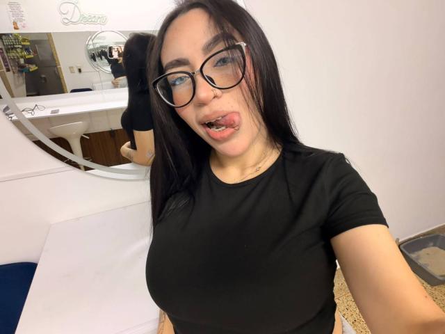Camille, "JuicyHot", met zwarte bril, lang zwart haar en haar tong uit, draagt een strak zwart shirt en laat haar grote borsten zien, op een selfie in een badkamer..
