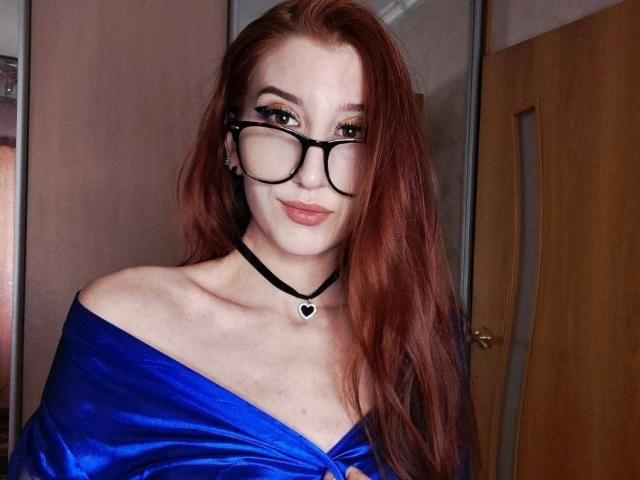 Edwina Torgerson hat rotes Haar, eine schwarze Brille, eine schwarze Halskette mit einem Herz und ein blaues Oberteil mit einem Schulterauschnitt, das ihre Brüste zeigt. Sie lächelt leicht und hat einen selbstbewussten Ausdruck..