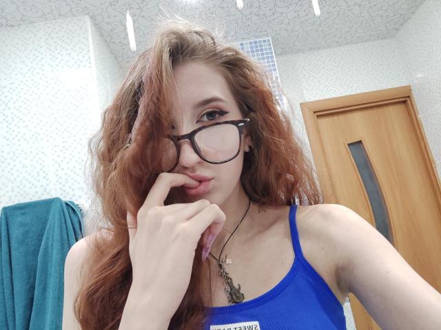 Edwina Torgerson, con lunghi capelli rossi, occhiali, canotta blu, dito vicino alla bocca, collana, pelle chiara, che guarda la fotocamera, porta di legno, asciugamano blu, pareti bianche con motivi, luci fluorescenti a soffitto..