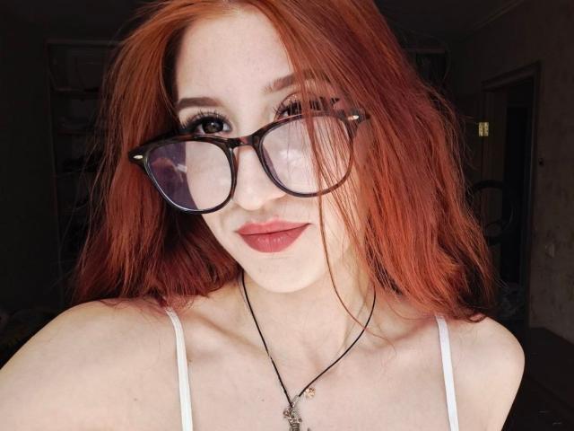 Edwina Torgerson, con cabello rojo, gafas y lápiz labial rosa, vestida con una blusa blanca de tirantes finos, un collar con un pequeño dije, sonriendo a la cámara..