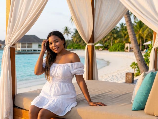 XinemaBonet, con piel morena, cabello castaño oscuro, pechos pequeños, vestida con un vestido blanco de un solo hombro, está sentada en una cama de playa, con la mano en el cabello, sonriendo. Se ven una playa y palmeras al fondo..