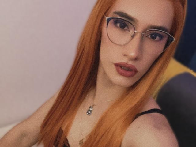CamiiNiieves, con lunghi capelli arancioni, occhiali e rossetto rosso, mostra il suo seno in una canottiera nera con spalline, indossando una collana con un piccolo ciondolo..