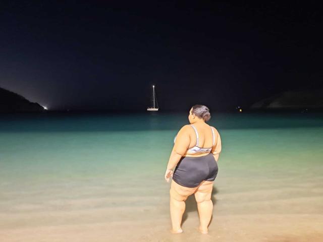 HurrenMatt, a fat woman with a large ass and big tits, wears a blue bikini, stands in shallow water, facing a dark night sea with a small sailboat.