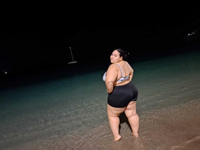 HurrenMatt has a big ass, large tits, and a curvy body, standing in shallow water at night, wearing a grey bra and black shorts, with dark hair in a bun.