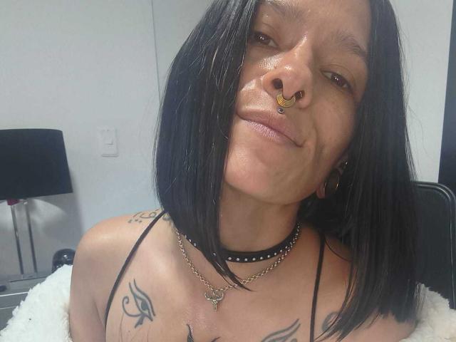 AmelieBlake with black bob, nose ring, choker, and tattoos, smiling slightly, showing her tits in a black strap top. White wall and lamp in the background.