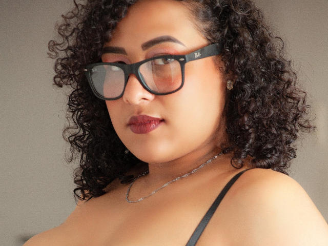 SamantaAngel mit lockigem, schwarzem Haar, die eine schwarze Brille, ein schwarzes Top mit Spaghetti-Trägern und roten Lippenstift trägt, hat eine silberne Halskette und einen leicht ernsten Gesichtsausdruck..