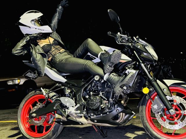 KimmyK, wearing black leather pants and jacket, sits on a black and white motorcycle with red wheels, legs up, arms raised, white helmet, black boots, dark night background.