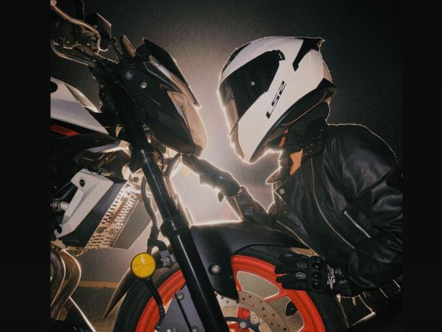 KimmyK trägt eine schwarze Lederjacke und einen weißen Helm mit einem schwarzen Visier und steht neben einem roten und schwarzen Motorrad, mit den Händen auf dem Lenker, in einer schwach beleuchteten Umgebung..