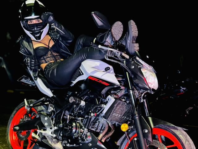 KimmyK, con un trasero grande, vestida de cuero y encaje negros, está sentada en una motocicleta blanca y negra con ruedas rojas, llevando un casco negro, con las piernas levantadas, por la noche..