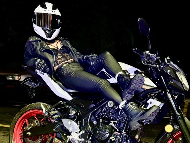 KimmyK trägt eine schwarze Lederjacke, ein Spitzenoberteil und enge Hosen und sitzt auf einem weißen Motorrad mit roten Rädern. Sie trägt einen weißen Helm. Im Hintergrund ist es dunkel..