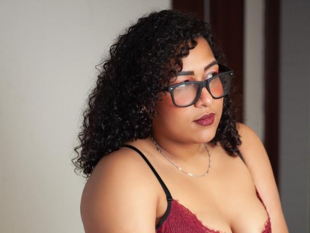 SamantaAngel, con i capelli neri ricci, gli occhiali e il rossetto rosso, indossa un reggiseno di pizzo rosso, guarda in basso con un leggero sorriso, ha una collana d'argento e un seno di media grandezza..