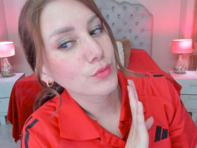 KatyaRogersX with pouty lips, wearing a red shirt, blue eyeshadow, and earrings, has a hand near her neck, pinkish-red room, and two lamps in the background.
