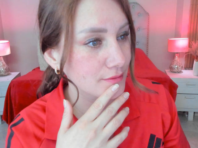 KatyaRogersX with pinkish skin, brown hair in a ponytail, wearing a red shirt, touching her chin, blue eyes, small earrings, slight smile, pinkish red bedroom lighting in background.