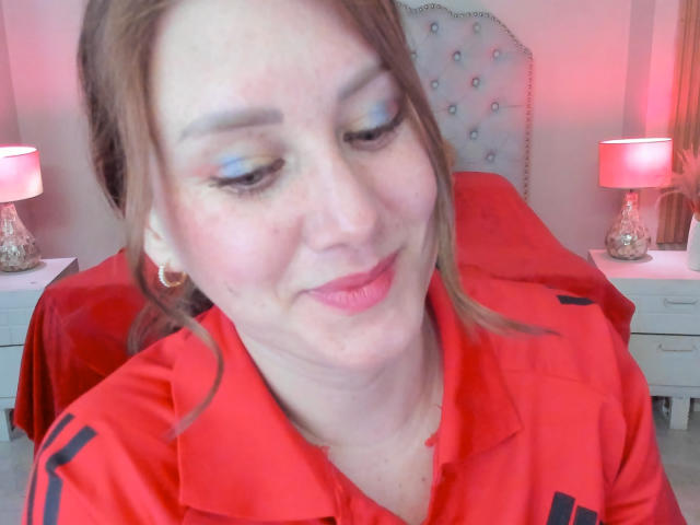 KatyaRogersX has light skin, red lipstick, blue and yellow eyeshadow, and brown hair in a ponytail. She's wearing a red shirt with black stripes, smiling.